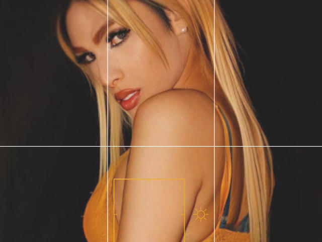 Putinna with long blonde hair, orange lipstick, and heavy eye makeup, looking over her shoulder, showing her bare back and yellow bra, with a sexy, sultry expression.