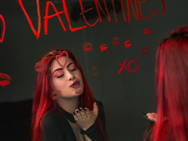 IsabellaRobertz, con capelli rossi, piercing al naso e ombretto rosa, labbra strette, mani vicino al viso, top nero, cuore rosso sulla fronte, smalto rosso, seconda persona con capelli rossi..