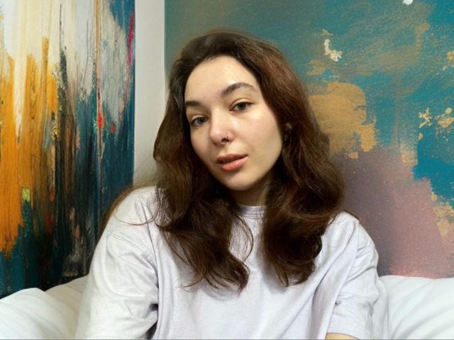 KissMegan with wavy brown hair, light skin, and pink lips, wearing a white shirt, sitting on a bed, looking at the camera with a relaxed expression.