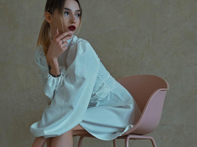 SophieKitty with blonde hair in a ponytail, wearing a white dress, sitting on a pink chair, hand near mouth, looking to the side, with red lipstick and a tattoo.