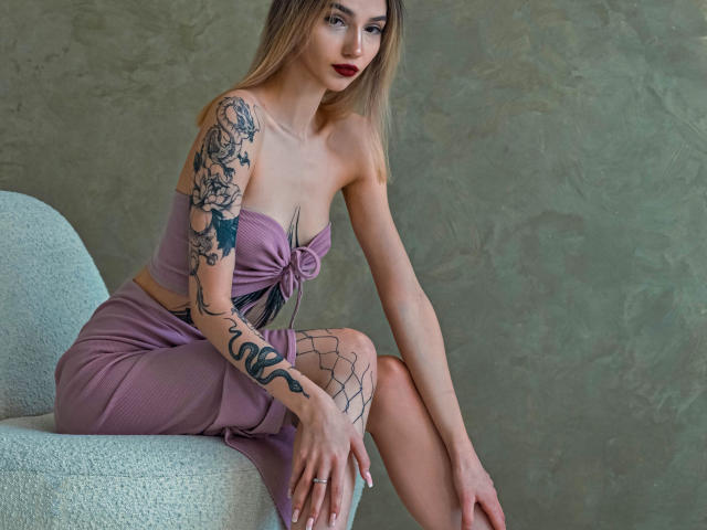 SophieKitty with blonde hair, red lipstick, and tattoos, wearing a purple strapless top and matching skirt, sitting on a white chair, showing her ass and small tits.