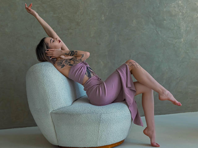 SophieKitty, com tatuagens, vestindo um vestido roxo e descalça, relaxando em uma cadeira branca, com os braços levantados, olhos fechados e pernas cruzadas, exibindo seu traseiro e seios pequenos..