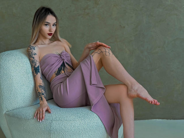 SophieKitty with tattoos, wearing a purple strapless dress, sitting on a white chair, legs crossed, showing her bare foot, and red lipstick, with blonde hair and dark eyes.