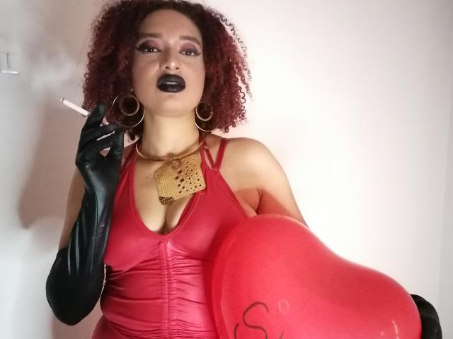 VioletaSwitch com cabelo ruivo cacheado, batom preto, vestido vermelho, luvas pretas, colar de ouro, segurando um cigarro, balão em formato de coração vermelho, brincos grandes em formato de argola, expressão confiante, pele clara..