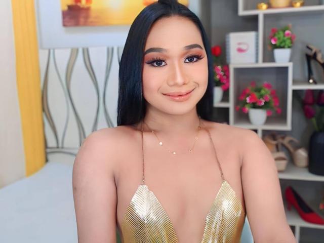 GiganticCummerx has long black hair, big brown eyes, and is wearing a gold, leaf-patterned, deep-cleavage top showing her tits. She's smiling with a nose piercing.