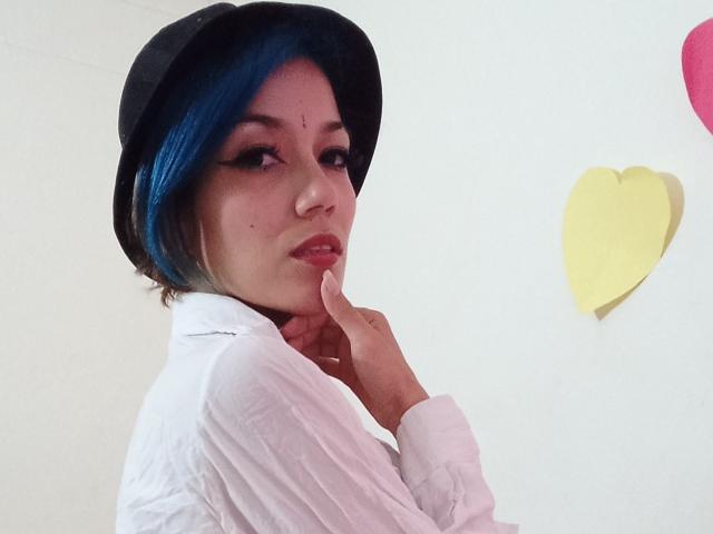 Veroo69 with blue-dyed hair, black hat, white shirt, and finger on chin, looking sexy. Yellow heart and pink heart in background. Soft light on her face.