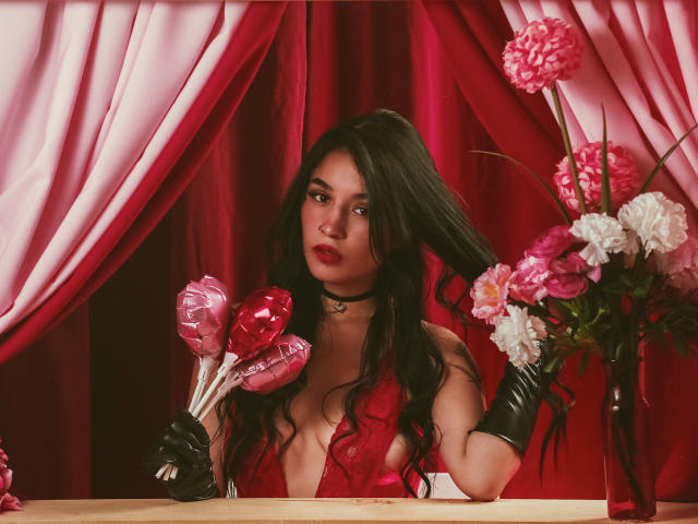 DulceHarper with dark hair, red dress, black choker, and gloves, holding pink heart-shaped candies, surrounded by pink flowers, red curtains, and a wooden table.