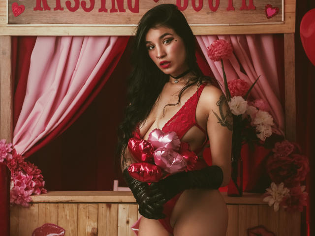 DulceHarper with long black hair, red lace lingerie, black gloves, holding pink ribbons, revealing her tits and ass, standing in front of a wooden booth with pink flowers.