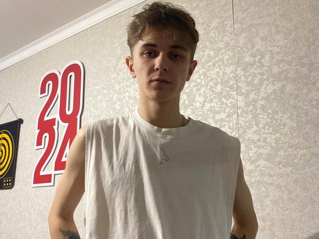 FranckyHotX, wearing a sleeveless white tee, with short brown hair and a serious expression, has a small tattoo on his right arm and a chain necklace.