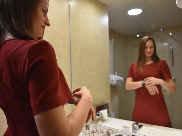 AbyX with tits in a red dress, looking in the mirror, adjusting her shirt with hands, reflection showing, in a bathroom, light beige walls, white sink, and mirror.
