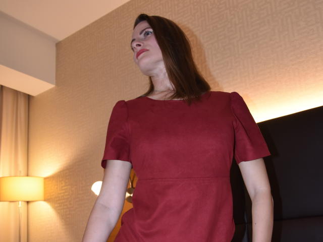 AbyX has pale skin, brown hair, and red dress with short sleeves, standing in a room, looking slightly up, with a lamp and curtain in the background.