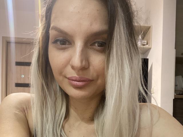 DouceAmelia has light brown skin, straight blonde hair with darker roots, and full pink lips. She's smiling softly, showing small, subtle freckles on her nose.