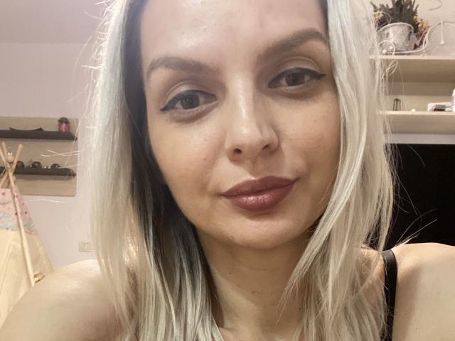 DouceAmelia has pale skin, blonde hair with blue highlights, black eyeliner, and pink lipstick. She's smiling with her tits barely visible in a black top.