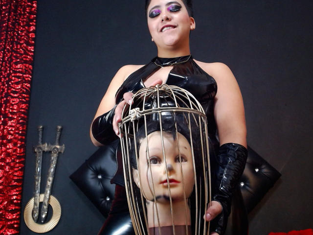 SamyWalker with dark makeup, black latex outfit, and gloves, holding a mannequin head in a birdcage, smiling. Mannequin has red lips, black head, and metal bars.