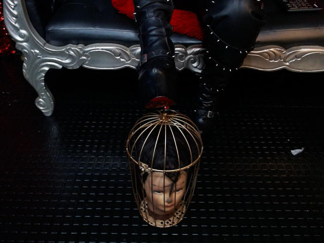 SamyWalker's black boot is kicking a gold birdcage with a man's face, black leather pants, and a red pillow in the background on a black floor.