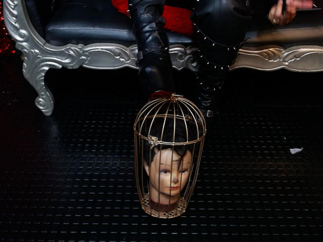 SamyWalker's face in a gold birdcage on a black floor, with black leather boots and a silver, ornate sofa in the background. SamyWalker's red shirt is partially visible.