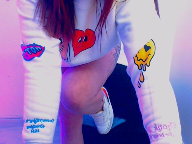 Kkatalina's white long-sleeve shirt with colorful heart and dripping yellow emoji tattoos, bending over, showing her white sneakers and bare ass. Her red hair falls forward.