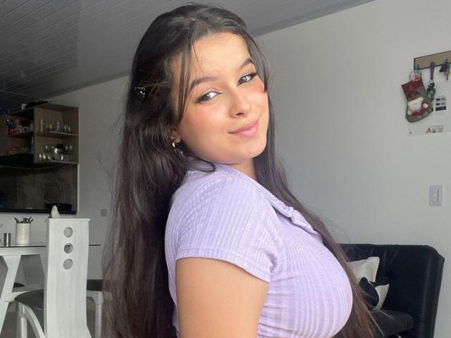 Kaary69 with long dark hair, wearing a purple ribbed top, smiling at the camera, showing her tits, in a bright room with white furniture.