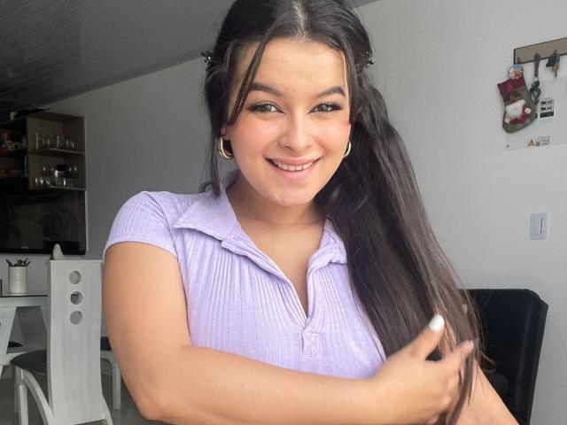 Kaary69 with long dark hair, smiling, wearing a purple ribbed shirt, gold earrings, holding her hair, showing her tits slightly, in a bright room.