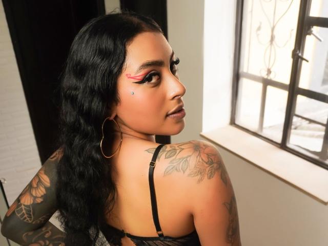 MagicMargo with wavy black hair, hoop earrings, pink eyeliner, tattoos on her right shoulder, and looking back. She's wearing a black lace bra, showing her back and ass.