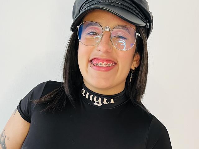 Natalie Harris con un berretto di pelle nera, occhiali trasparenti, maglia a collo alto nera con colletto a punte, camicia nera e denti con "griglie" visibili, sorridente, capelli neri, sfondo bianco, tatuaggio sul braccio sinistro..