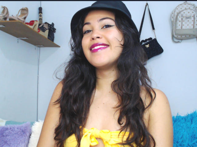 SharonBurtton has long wavy black hair, a black hat, yellow strapless top, and pink lipstick. She's smiling with visible tits, and has a cute, attractive face.
