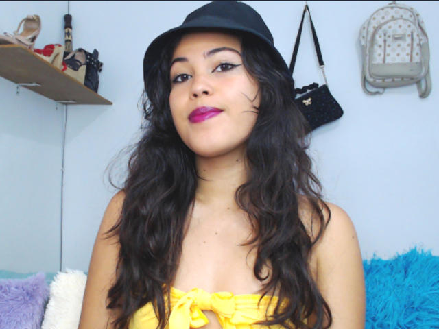 SharonBurtton with wavy black hair, wearing a black hat, yellow strapless top, and pink lipstick, has a cute face and small tits, standing in a room with colorful pillows.