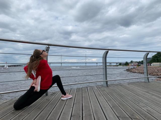 BiancaHotLove is on a wooden boardwalk, kneeling with her back to the camera, wearing a red sweater, black pants, and pink sneakers, stretching her ass upwards.