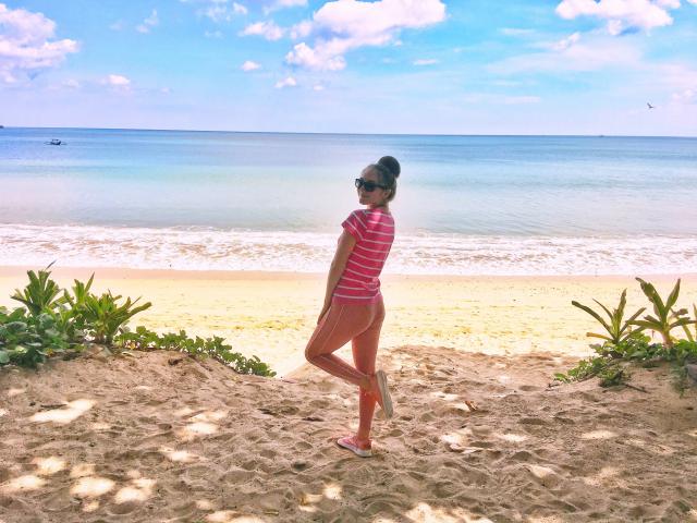 BiancaHotLove with her ass in pink striped shirt and orange pants, standing on sandy beach, looking back, wearing sunglasses, and her hair in a bun, near green plants.