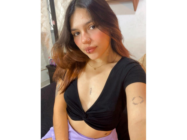 SophiaMarx with brown hair, wearing a black tied-up crop top, showing small tits, purple pants, a heart tattoo on her right arm, and a small chest tattoo.