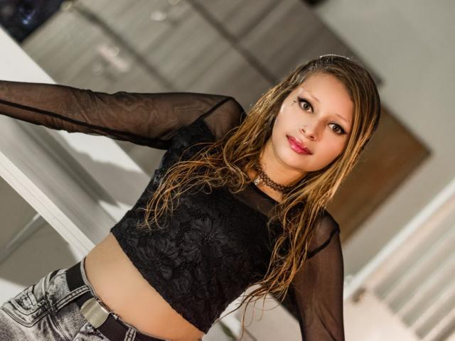 KeiliSexy with wet blonde hair, black lace top, sheer sleeves, and choker, standing, showing her tits, wearing denim jeans, and looking at the camera with red lipstick.