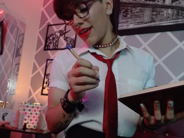 LinciSofia with glasses, red tie, white shirt, choker, holding pencil, tattooed arm, smiling, pink-red lighting, short dark hair, nose ring, wrist bracelet, nail polish, red lipstick.
