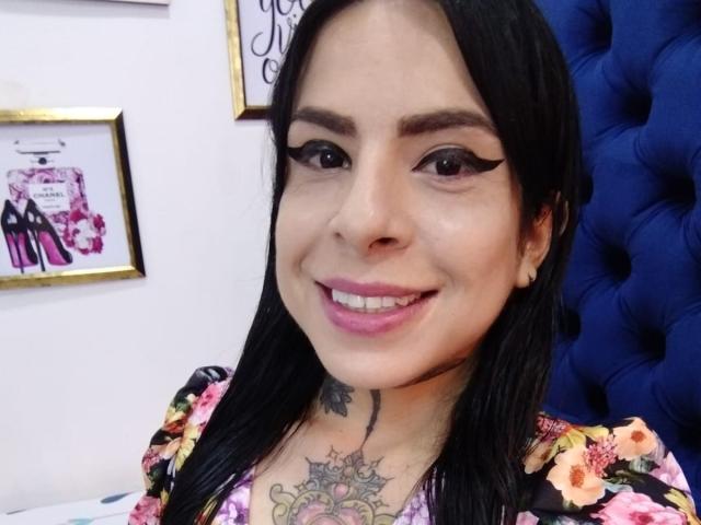SaoryPrincess has black hair, pink lipstick, and a floral dress with colorful flowers. She's smiling with winged eyeliner, showing tattoos on her chest and neck.