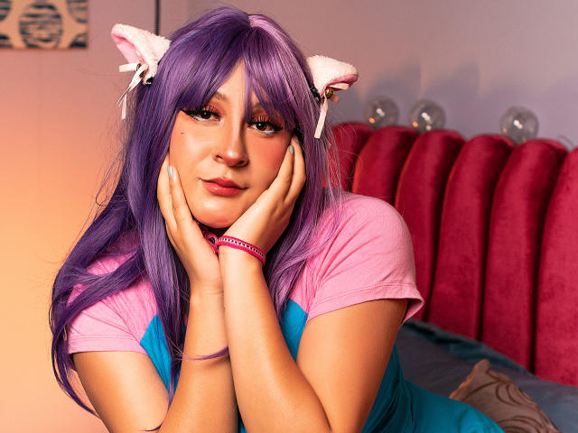 KittyEla mit lila Haaren, rosa Katzenohren und einer rosa Kette, die ihre Hände auf ihr Gesicht legt, trägt ein rosa und blaues Hemd und sieht süß und entspannt aus..