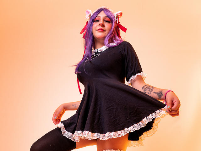 KittyEla com cabelo roxo, orelhas de gato, vestido preto com renda branca, segurando a saia, meias pretas até a coxa, tatuagens, fitas vermelhas, sorrindo, em pé, iluminação laranja suave..