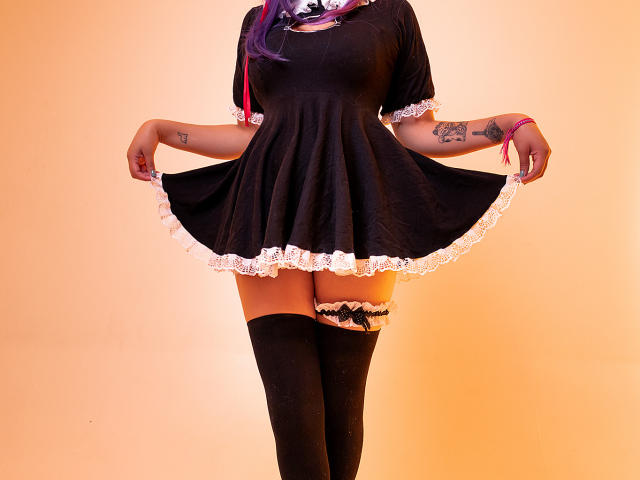 KittyEla vestindo um vestido preto com renda branca, segurando-o, mostrando meias pretas até a coxa com uma parte superior branca de renda, tatuagens nos braços, cabelo roxo e uma pulseira rosa..
