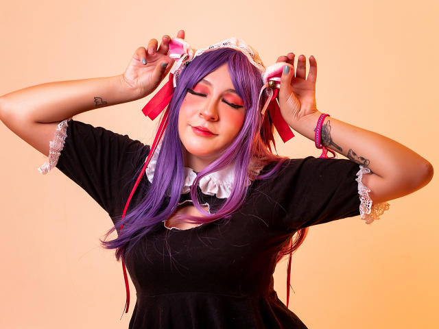 KittyEla com cabelo roxo, vestido preto, gola de renda branca e fitas vermelhas, segurando fitas, sorrindo, batom rosa, tatuagem no antebraço direito, pulseira rosa, luz quente, expressão relaxada..