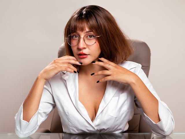 KittyEla com cabelo curto castanho, óculos, camisa branca, unhas pretas, mãos perto do rosto, decote discreto, olhando para a câmera, sentada, mesa de vidro, iluminação suave, expressão neutra..