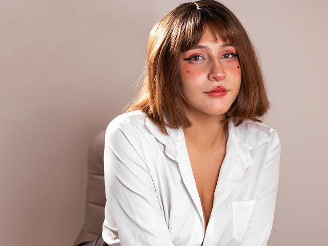 KittyEla with shoulder-length brown hair, white shirt, pink eyeliner, and black nose dots, sitting against a plain wall, looking slightly to the side with a subtle smile.