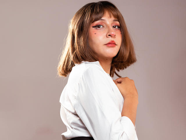 KittyEla, avec une coupe de cheveux courte marron, une chemise blanche, un eye-liner rose et des points rouges sur le nez, regardant par-dessus son épaule, une main sur la poitrine, fond gris clair..