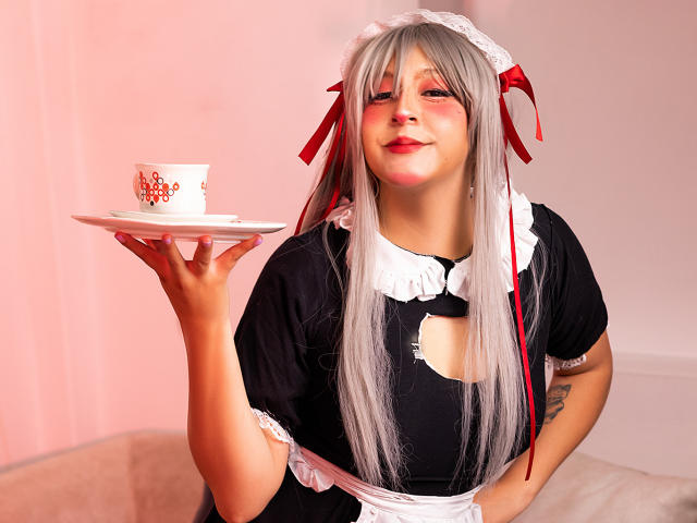 KittyEla, com longos cabelos prateados, vestindo um uniforme de empregada preto, segurando uma xícara branca com padrões vermelhos em um pires, sorrindo com lábios rosados. Seus seios são pequenos..