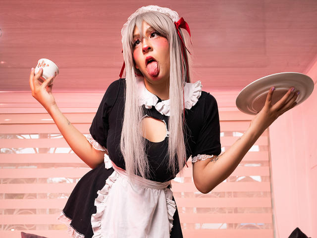 KittyEla in a black maid outfit with white frills, holding a egg in one hand and a plate in the other, tongue out, gray hair, red ribbon, looking crazy.