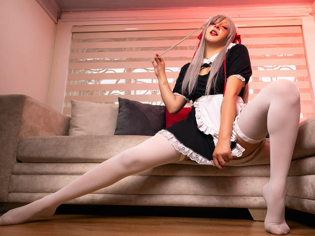 KittyEla with long silver hair, black and white maid outfit, white thigh-highs, and red ribbon, sitting on couch, legs spread, holding string, red light, sexy, cute, confident.