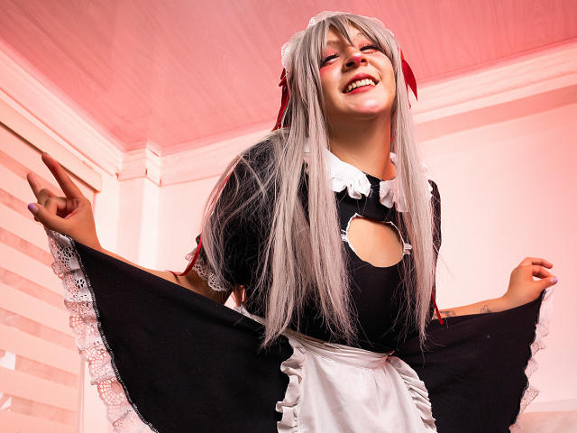 KittyEla, con cabello plateado largo, ojos rojos y un uniforme de sirvienta negro, sonriendo, extendiendo su falda, mostrando un corte en su pecho, en una habitación iluminada con luz rosa..