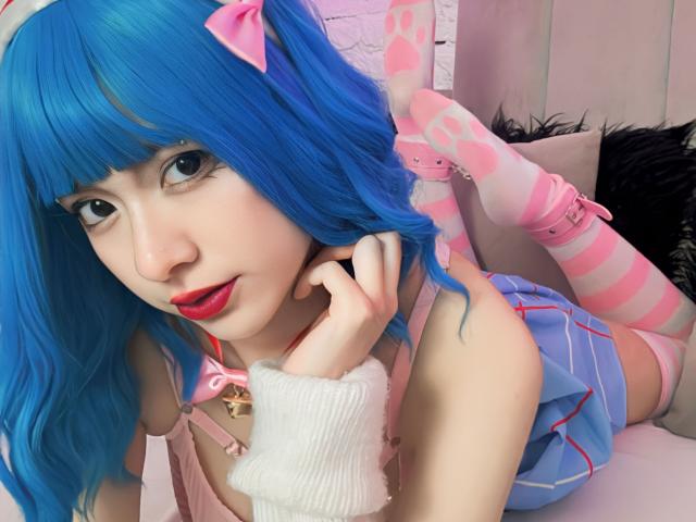 SabriPrettty with blue hair, pink bow, red lipstick, and striped pink socks, lying on a bed, wearing a pink harness and blue skirt, showing her tits and ass.