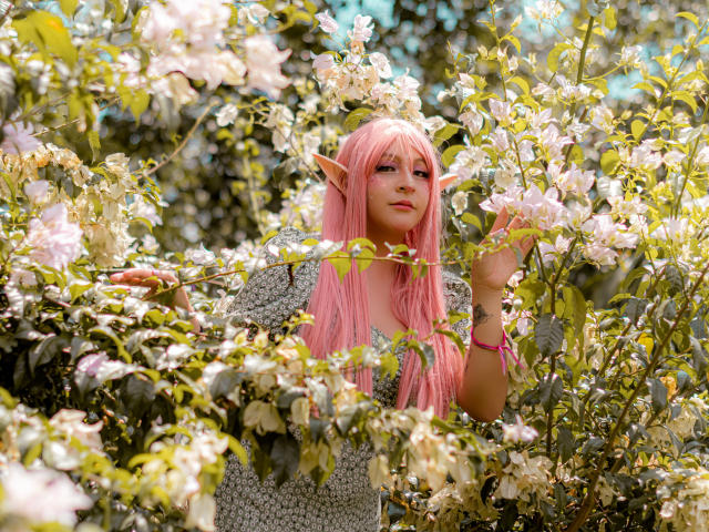 KittyEla, con cabello rosa, orejas de elfo y un vestido gris con estampado, rodeada de flores blancas en flor, sosteniendo una rama, luciendo linda y mágica..