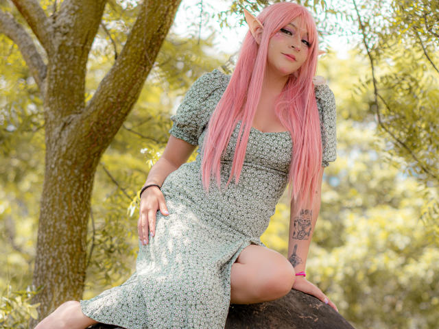 KittyEla, con cabello rosa, orejas de elfo y un vestido floral, sentada en un árbol, mostrando su brazo y muslo tatuados, con una sonrisa sensual. Fondo de bosque..