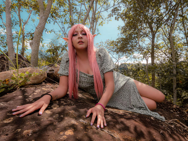 KittyEla, con orecchie da elfo rosa, lunghi capelli rosa e un vestito grigio, sdraiata su una roccia in una foresta, con le mani e i glutei a terra, che guarda verso la fotocamera..
