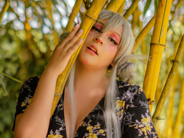 KittyEla, com cabelo grisalho, maquiagem rosa nos olhos e uma camisa florida, segurando bambu amarelo. Ela está olhando para cima com um leve sorriso, mostrando sardas e lábios rosados..