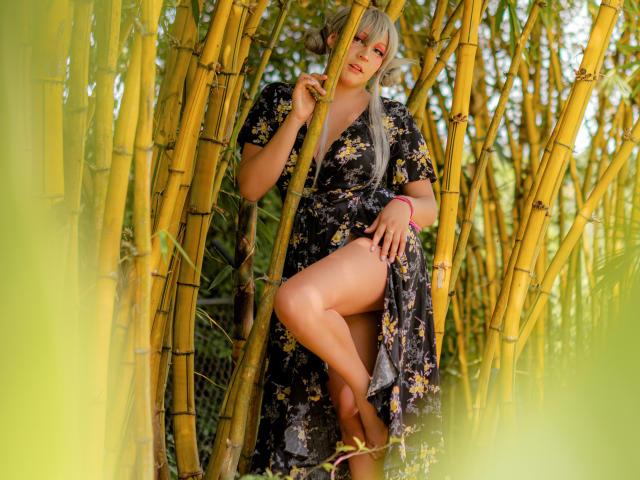 KittyEla mit blonden Haaren, einem Blumenkleid und einem rosa Armband, steht inmitten von Bambus, hält einen Stock, zeigt ein Bein, schaut in die Kamera, sonnig, gelber Bambus, sexy, süß..